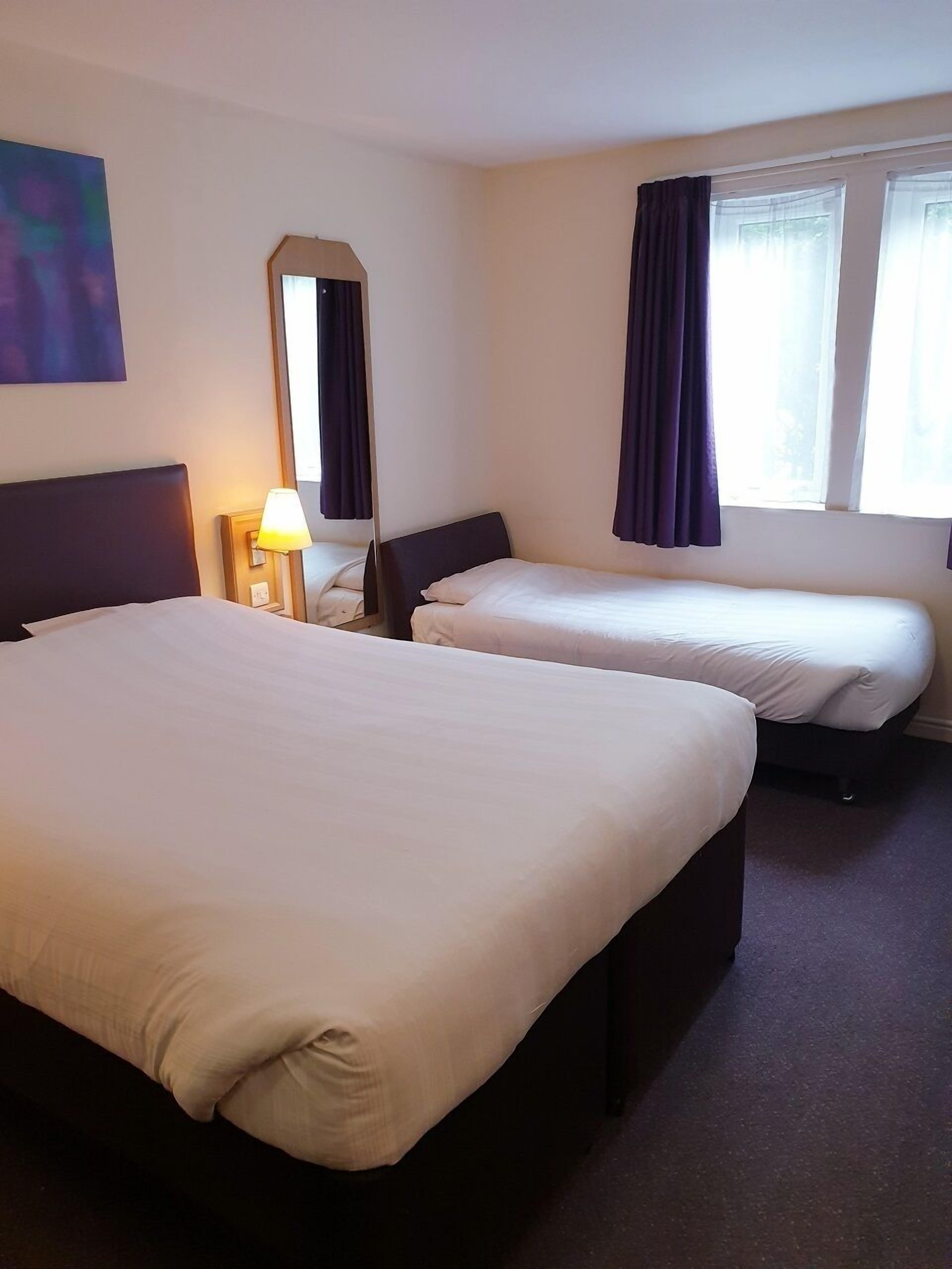 A hotel room with two beds, a nightstand with a lamp, a large mirror, and a window with curtains.