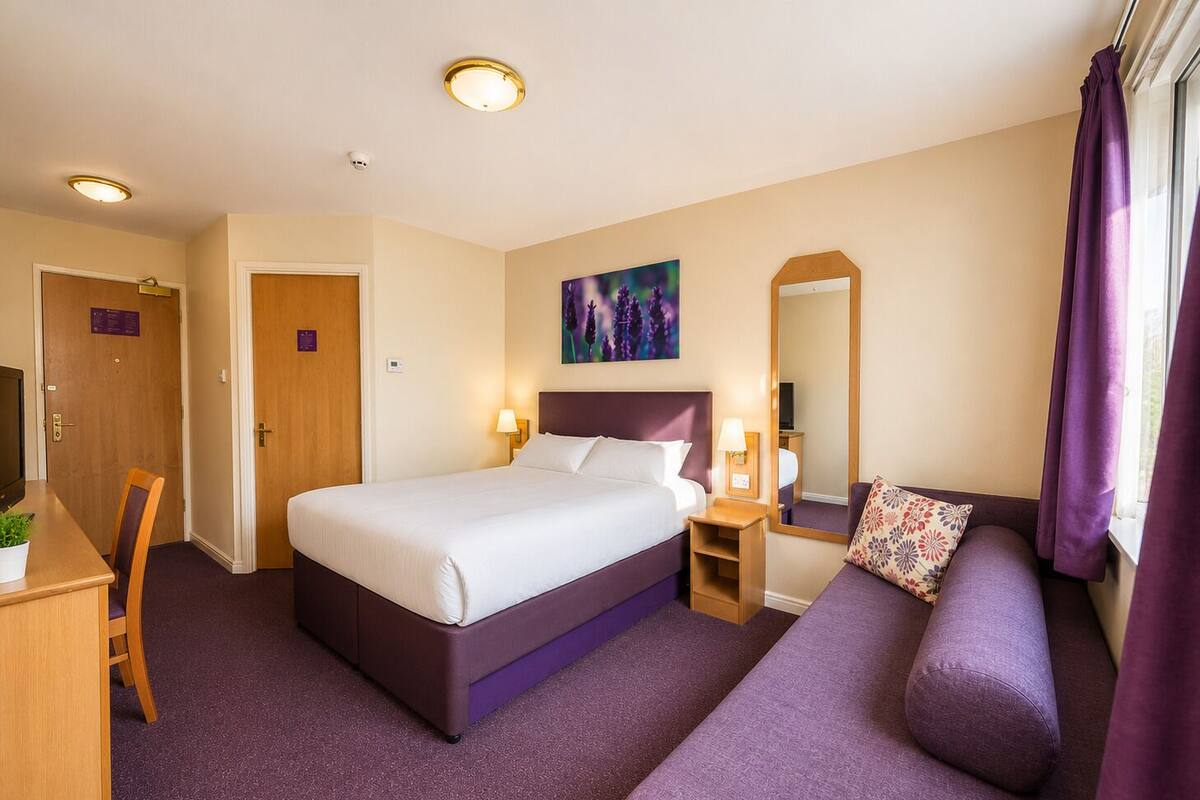 A hotel room with a bed, bedside tables, a sofa, a desk with a chair, a TV, and a window with curtains.