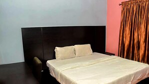 A bedroom with a large bed, a dark headboard, a small table, and a curtain. - D'ISLAND hotel (Lekki)