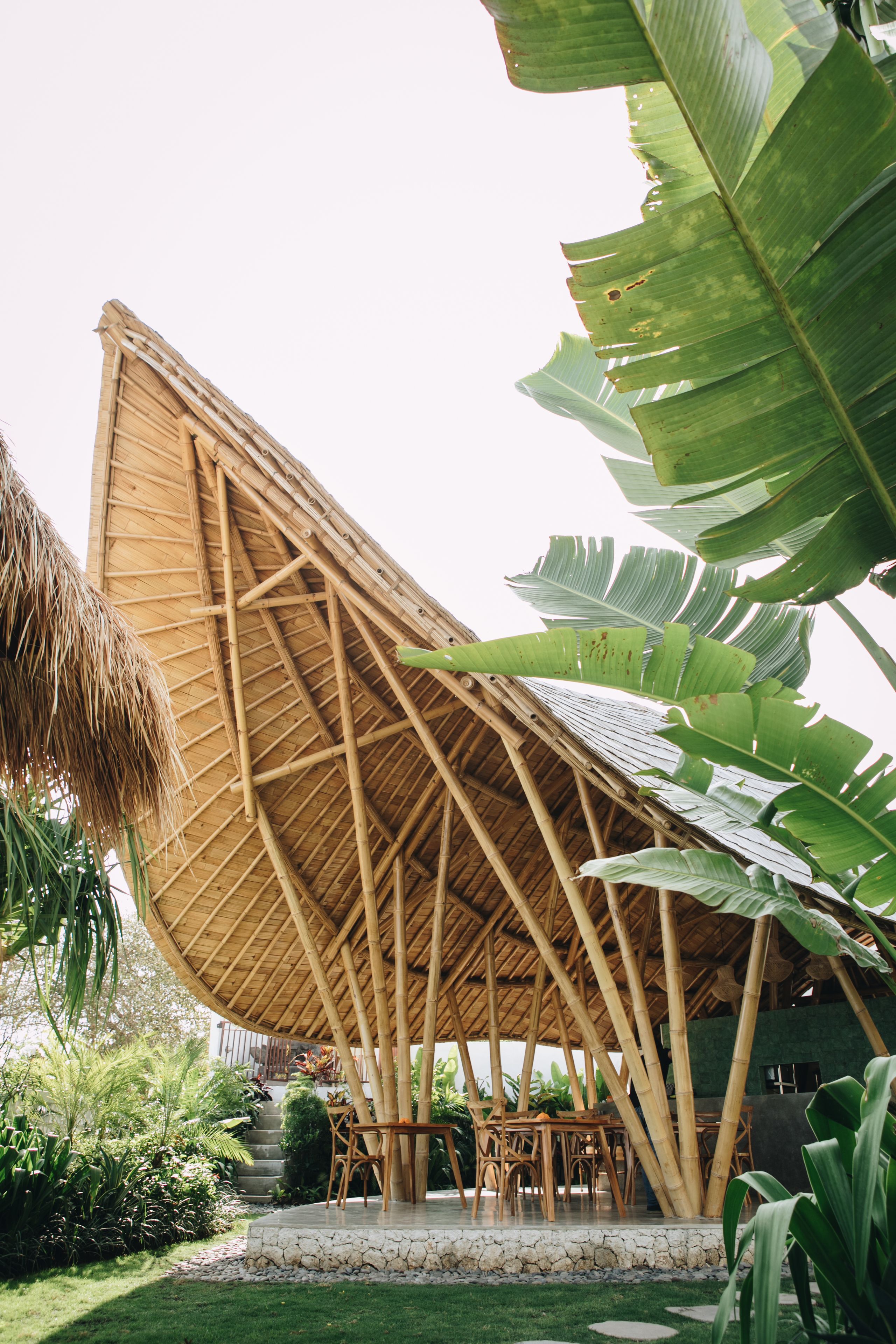 A bamboo structure with a thatched roof, surrounded by lush greenery and a stone pathway.
