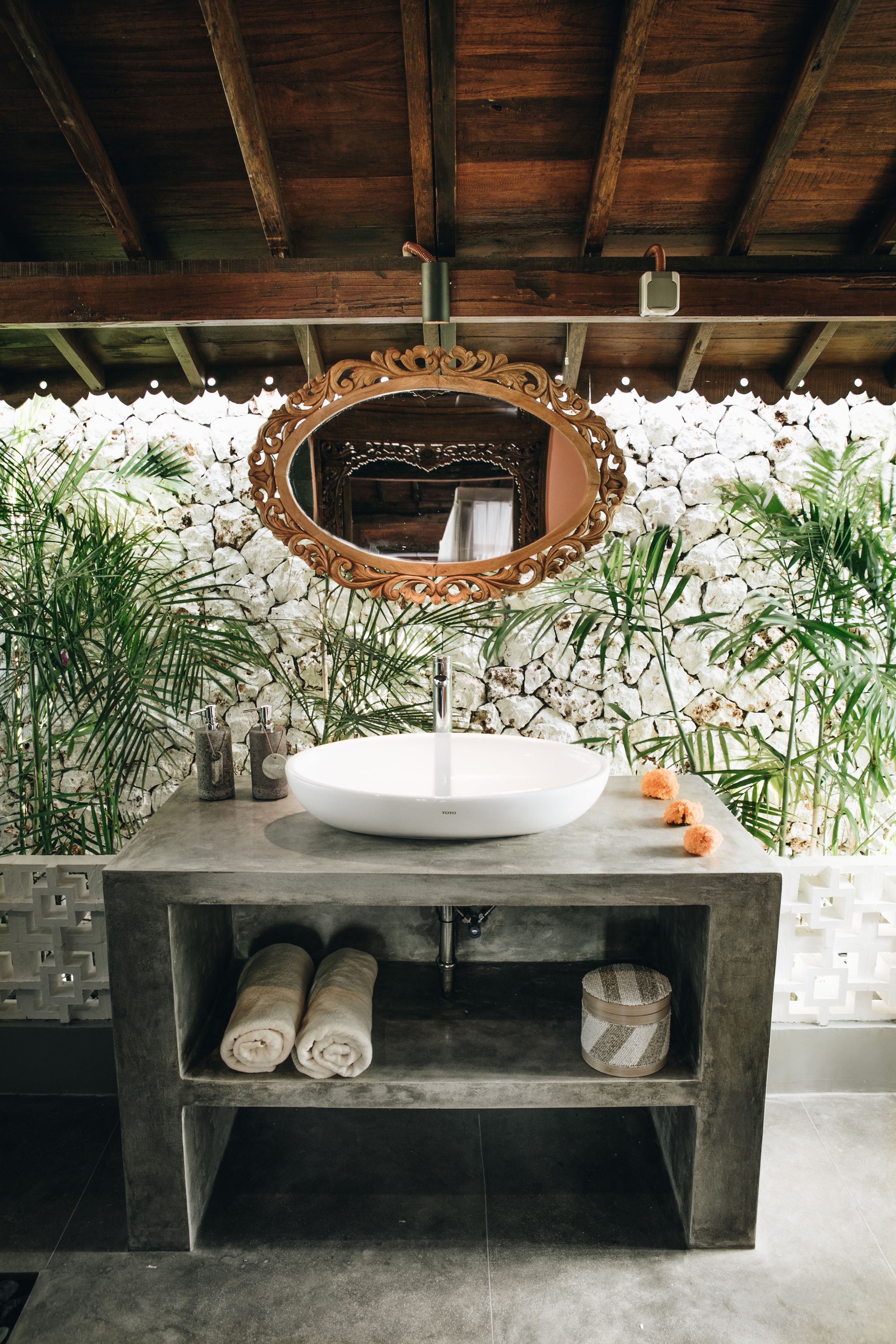 A bathroom with a wooden ceiling, a white sink, a decorative mirror, and a stone wall.