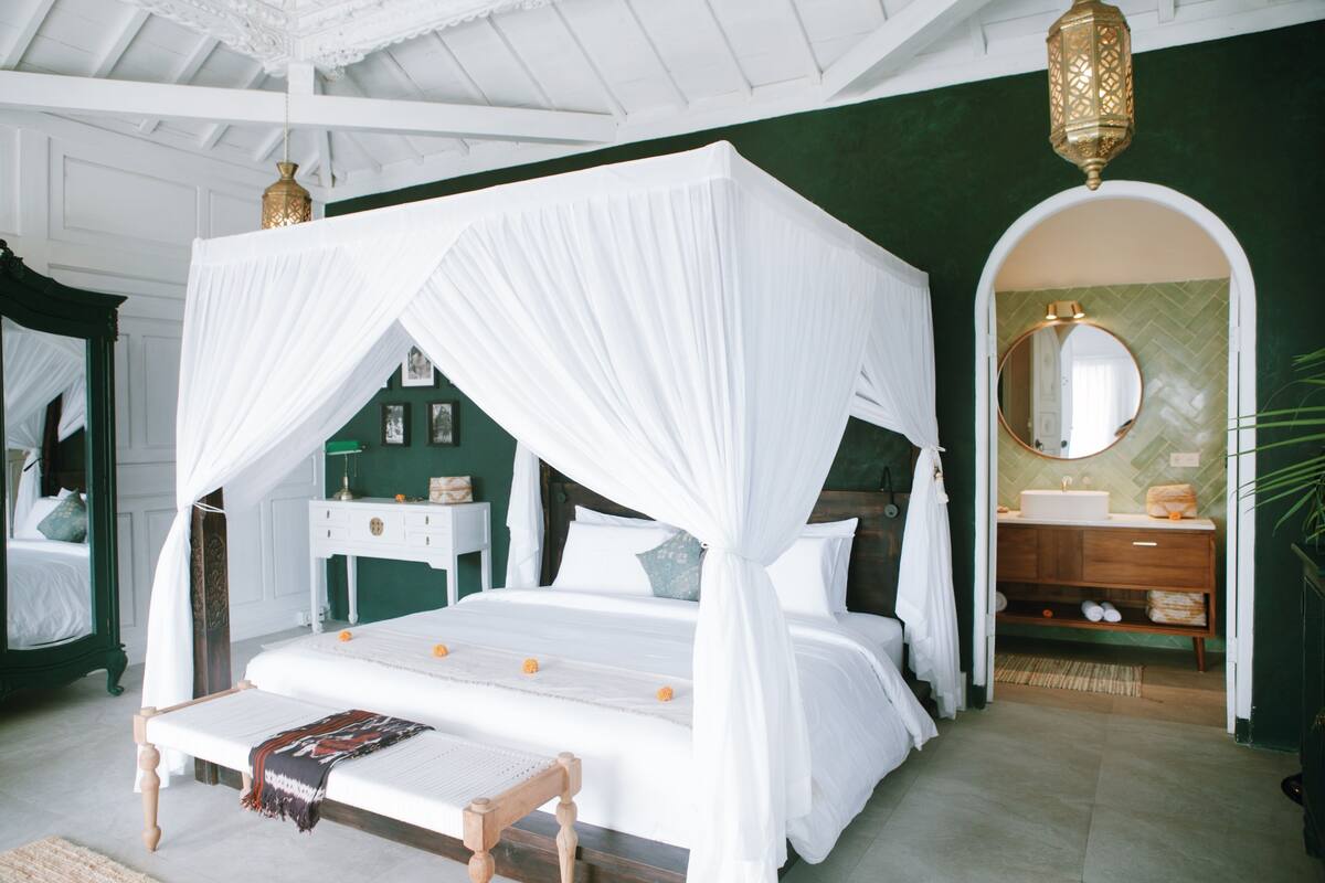 A bedroom with a four-poster bed, a bedside table, a mirror, and a bathroom with a sink and vanity.