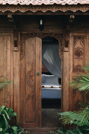 A wooden door with intricate carvings, a bed with a white mattress and a dark blanket, and a small plant to the left. - White Tortoise Eco Villa's (Pecatu)