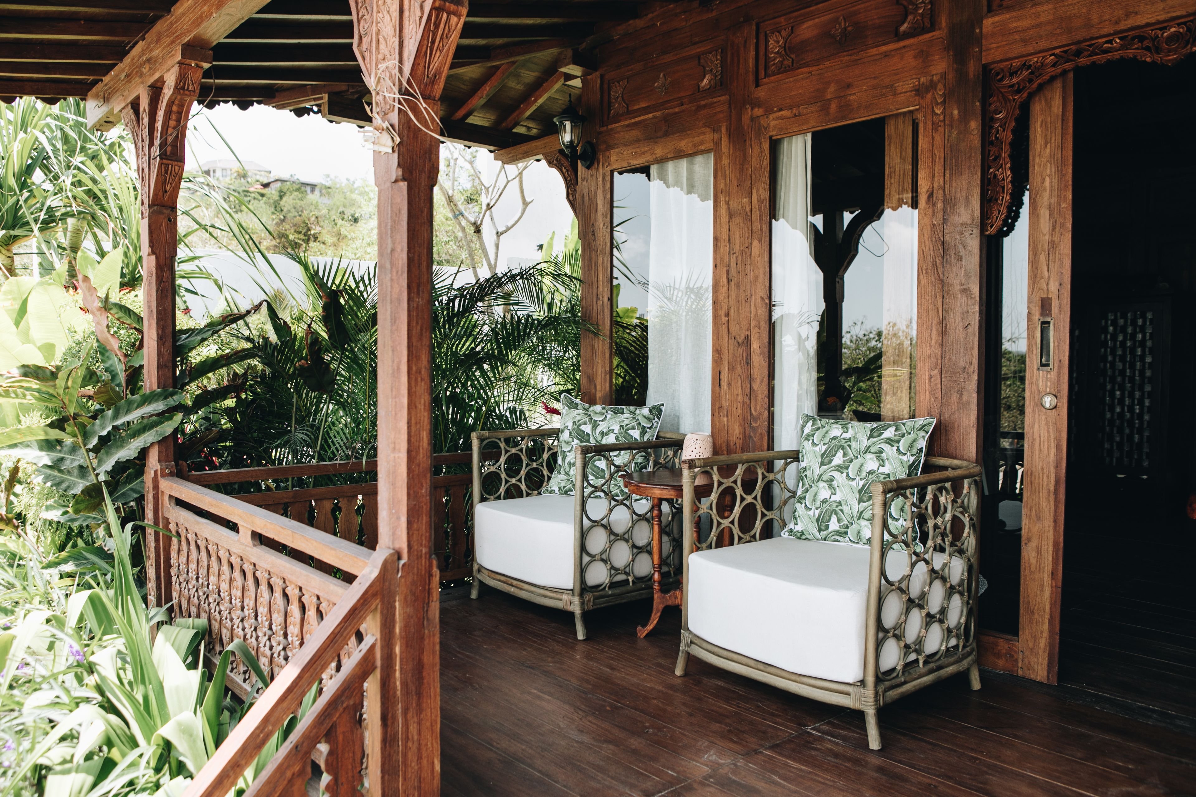 A wooden porch with two wicker chairs and a small table, surrounded by lush greenery.