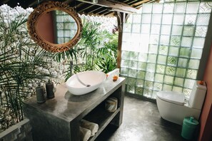 A bathroom with a large mirror, a white sink, and a toilet. There are plants and a window with a decorative glass pattern. - White Tortoise Eco Villa's (Pecatu)