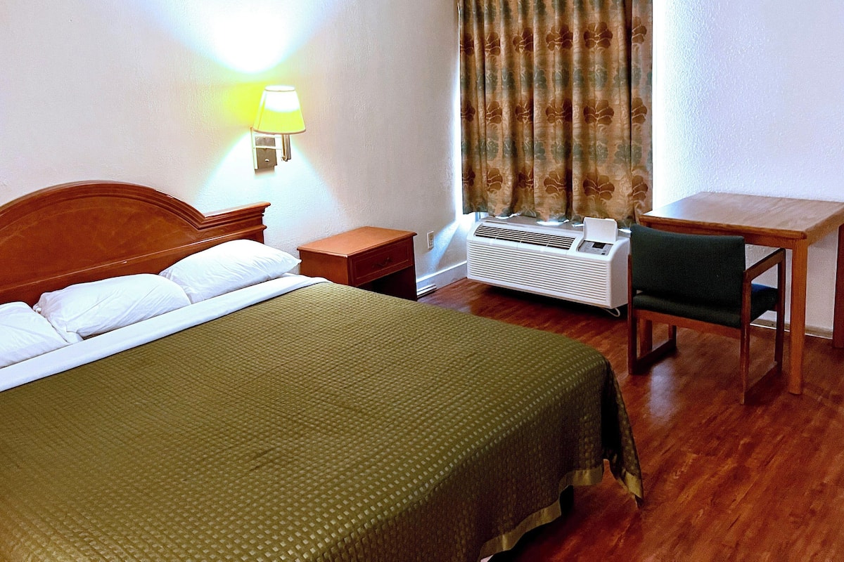 A hotel room with a bed, bedside table, lamp, air conditioning unit, and a small desk with a chair.