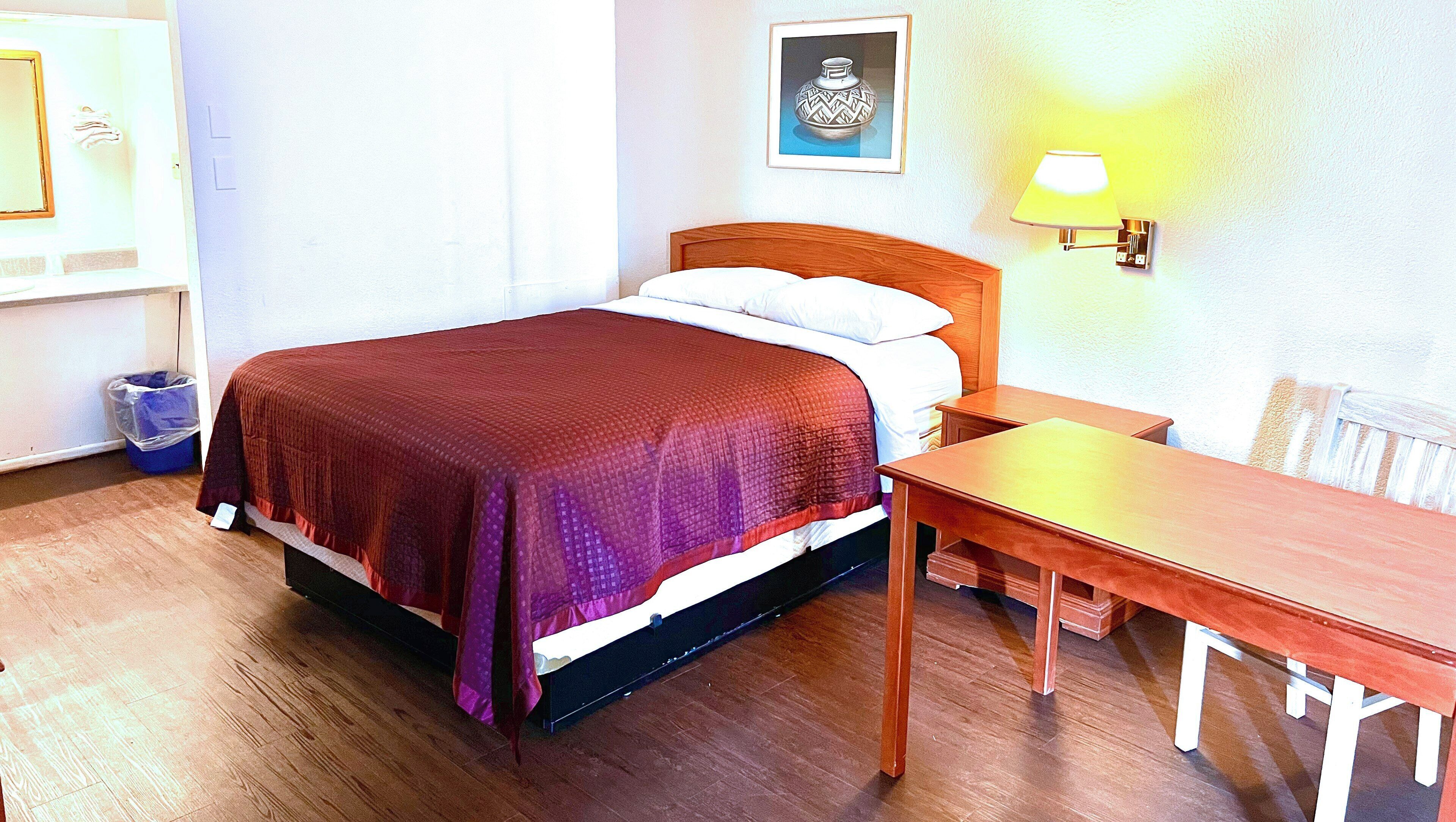 A room with a bed, a wooden nightstand, a table, a trash bin, and a wall-mounted lamp.