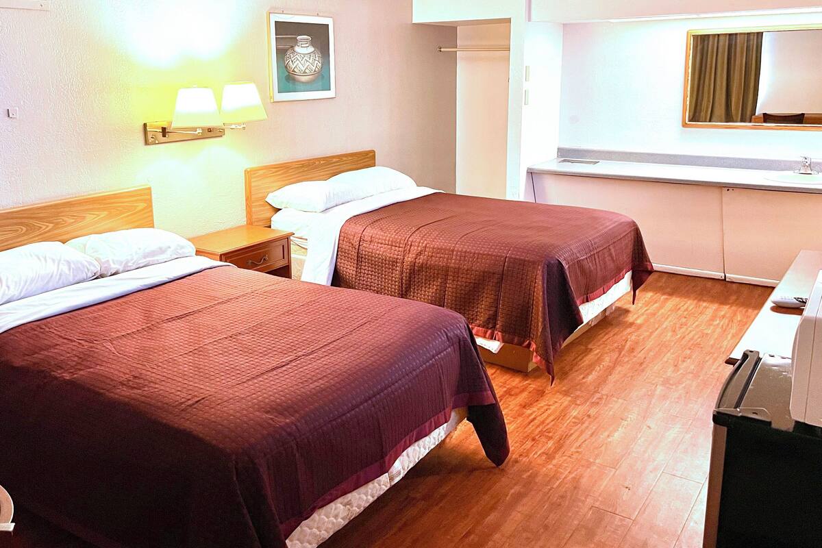 A hotel room with two beds, a nightstand, a desk with a chair, a mirror, and a wall-mounted lamp.