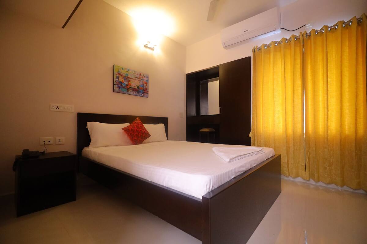 Hotelkamer met een bed, nachtkastje, airconditioning aan de muur en een raam met gele gordijnen.