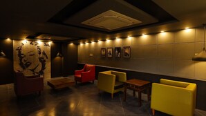 A modern lounge area with a high ceiling, recessed lighting, and a central air vent. There are several seating options including a red chair, yellow armchairs, and a wooden table. The walls feature framed artwork and a large abstract mural. - Kfour Apartment & Hotels Private Limited (Madurai)