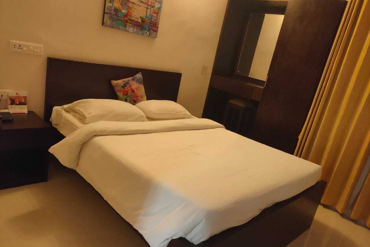 Een netjes opgemaakt bed met witte lakens, een donker hoofdbord, een kleurrijk kussen, een nachtkastje met een lamp en een spiegel aan de muur.
