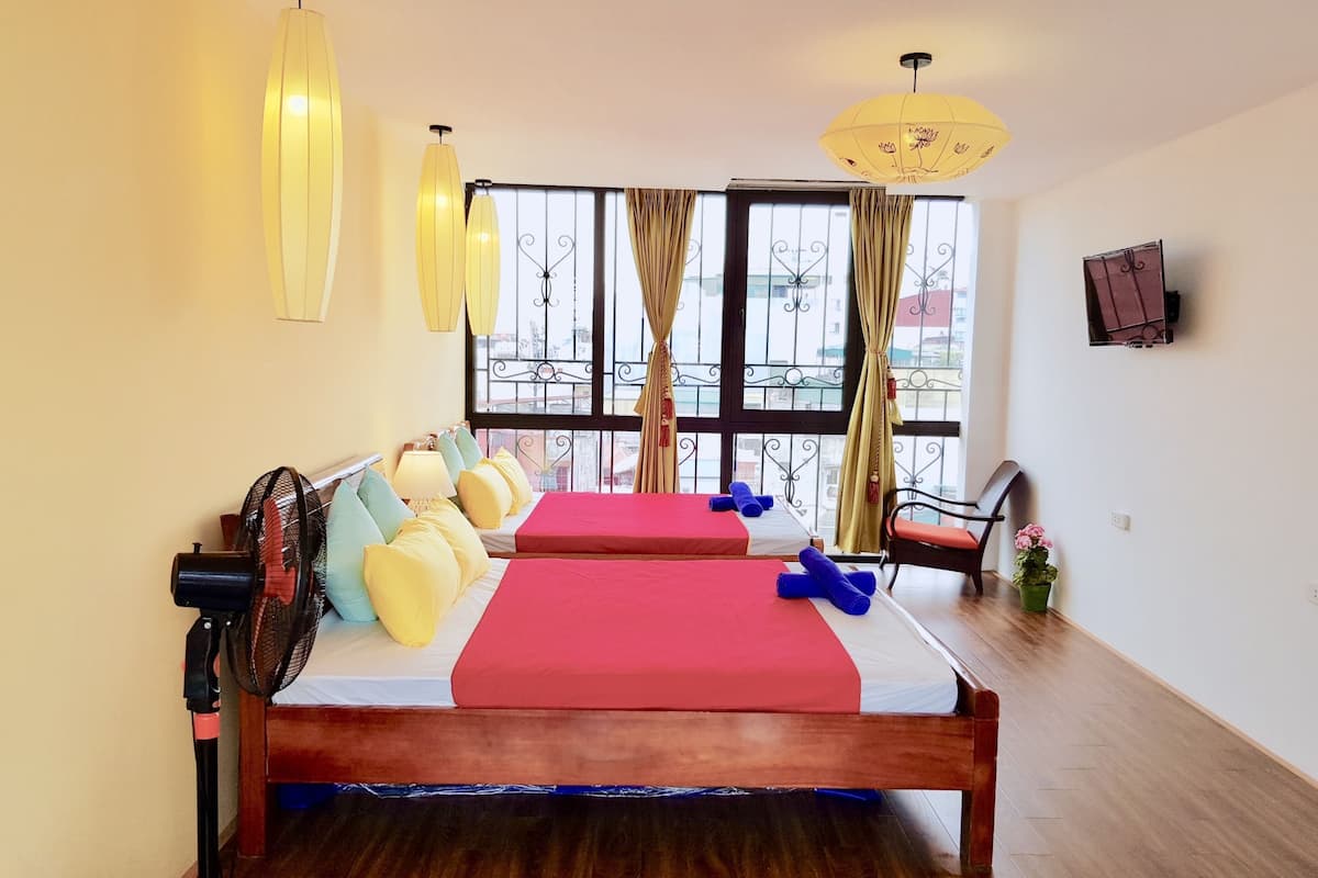 Ein Hotelzimmer mit zwei Betten, Holzboden, großem Fenster mit Vorhängen, einem Standventilator und einem angebrachten Fernseher.