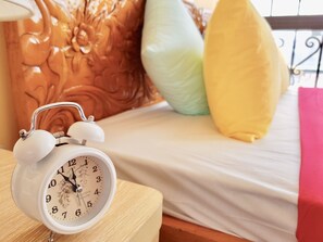 A bed with a white sheet, a wooden nightstand with a white alarm clock, and decorative pillows in orange, green, and yellow.