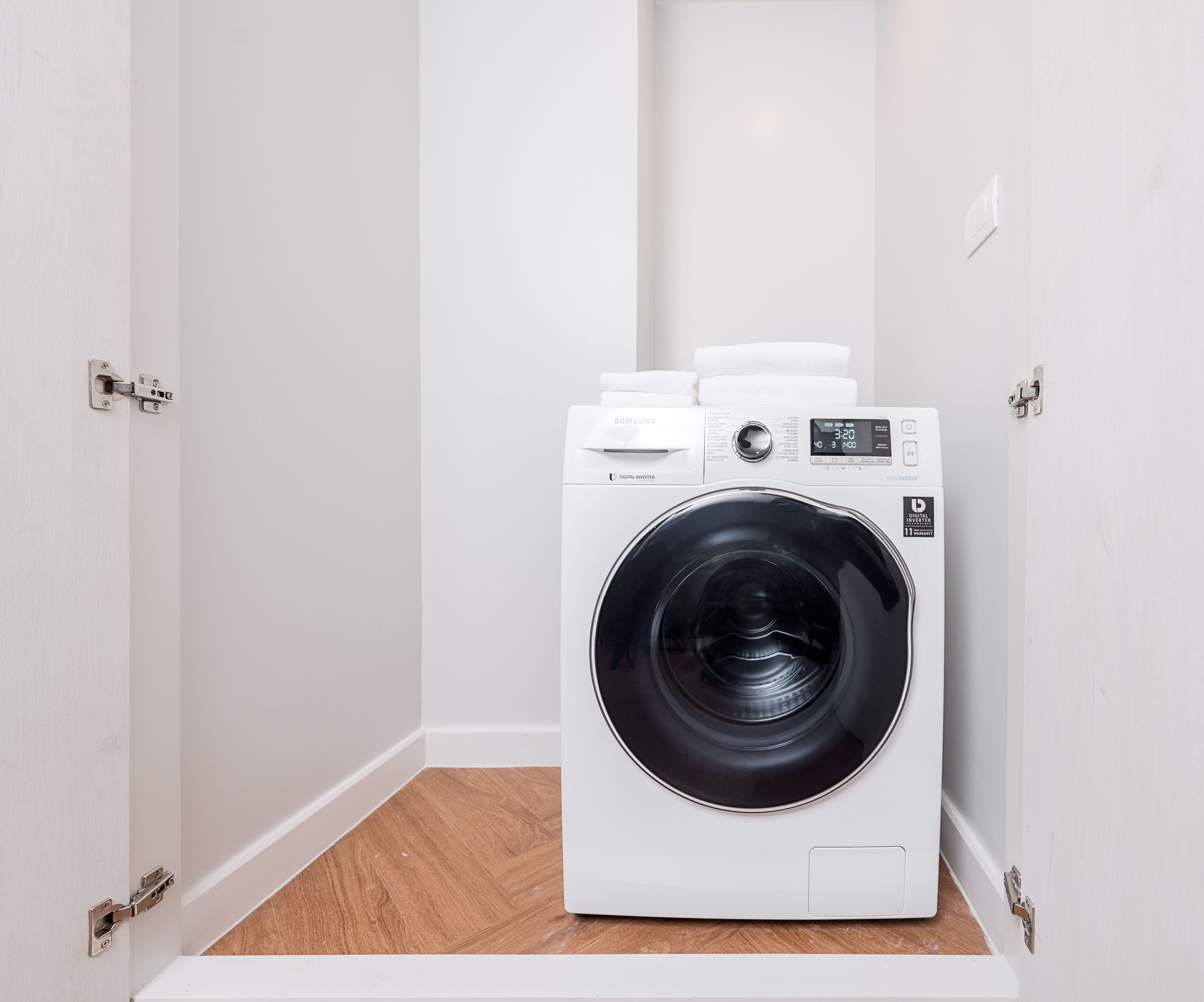 A compact, white front-loading washing machine in a small laundry room with wooden flooring and white walls.