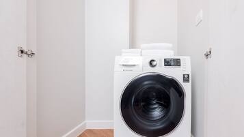 A compact, white front-loading washing machine in a small laundry room with wooden flooring and white walls.