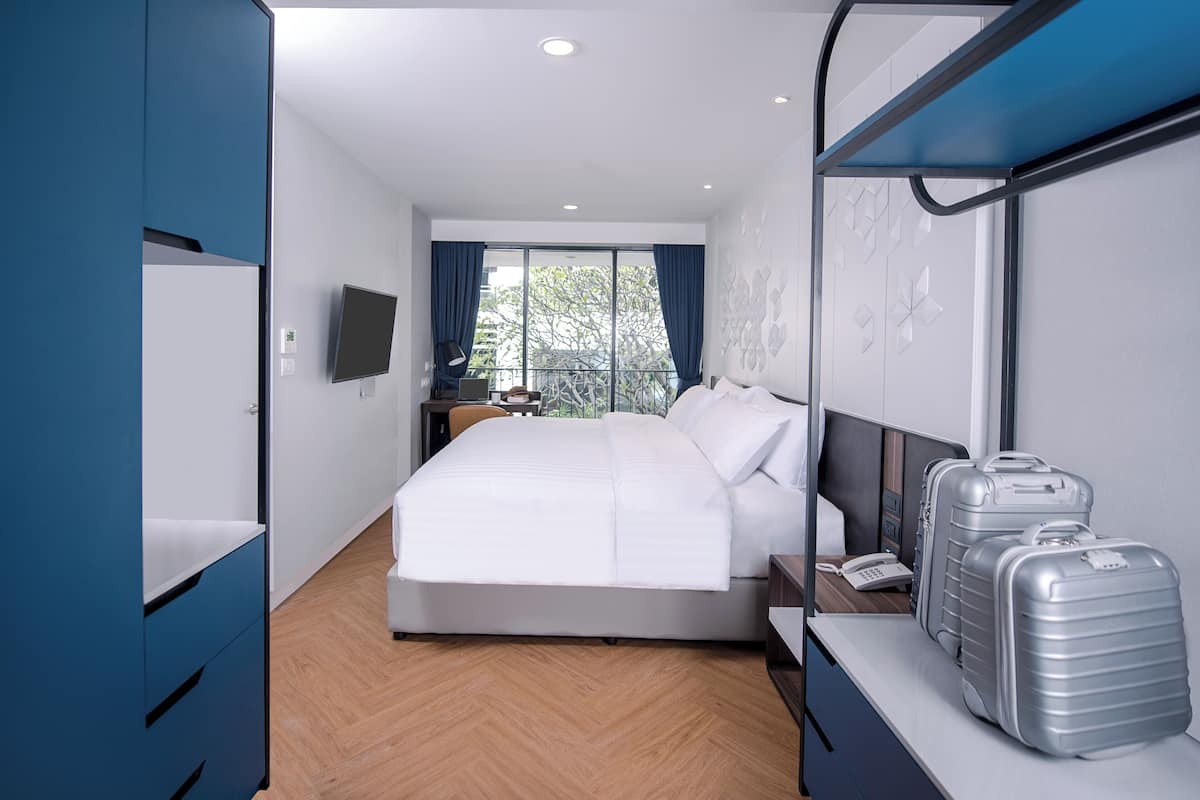 Una habitación de hotel moderna con una cama grande, un escritorio con computadora, un televisor de pantalla plana y un balcón con vistas a árboles.