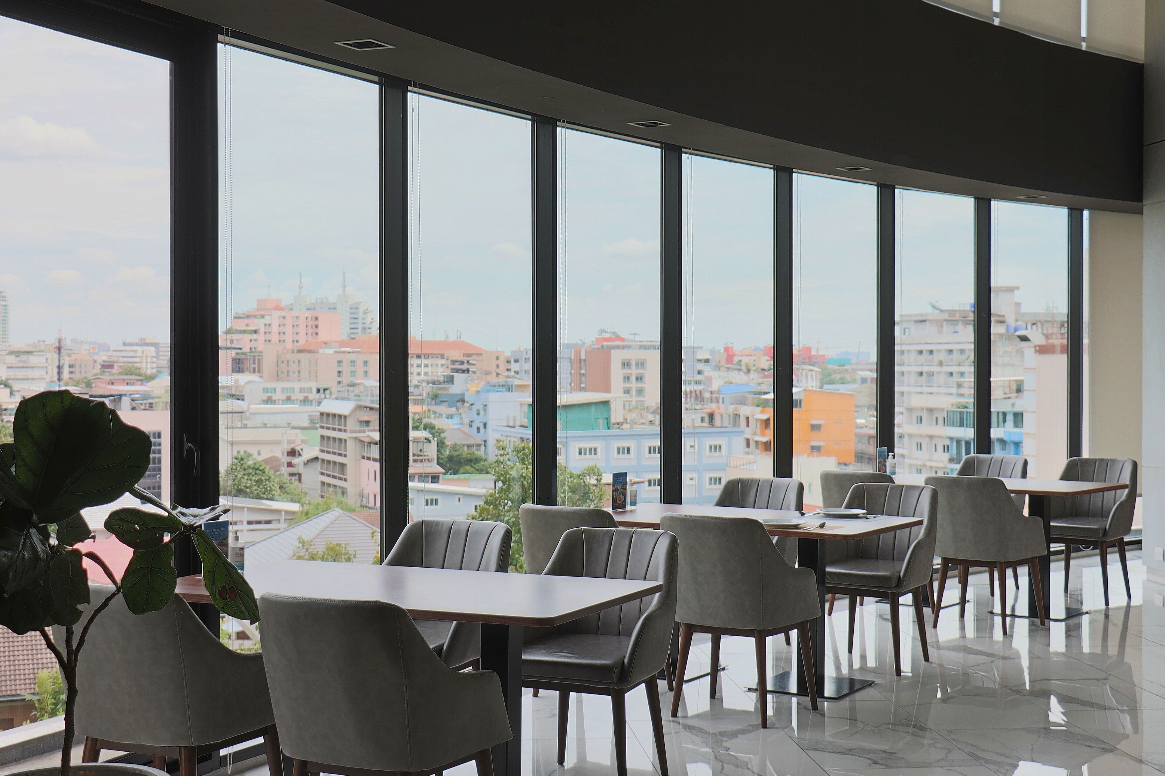 Un comedor moderno con ventanas amplias, varias mesas y sillas, que ofrece vista a un paisaje urbano.