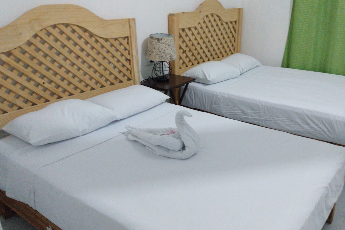 A hotel room with two single beds, wooden headboards, white linens, a swan-shaped towel, a lamp, and a green curtain.