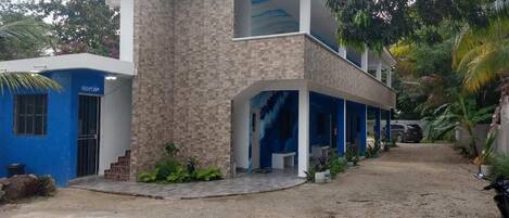 Un edificio de dos plantas con fachada azul y beige, un espacio al aire libre cubierto con asientos y un camino de entrada de grava.