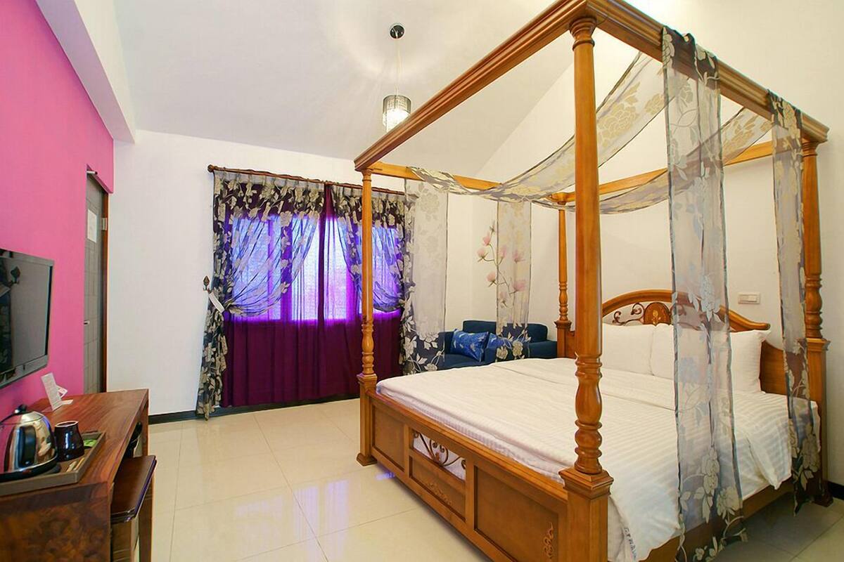 A four-poster bed with a canopy, a television on a wooden desk, and a purple curtain.