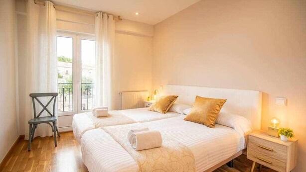 Una cama doble con cabecera blanca, dos mesitas de noche con lámparas, una silla y vistas al exterior a través de una ventana con cortinas.