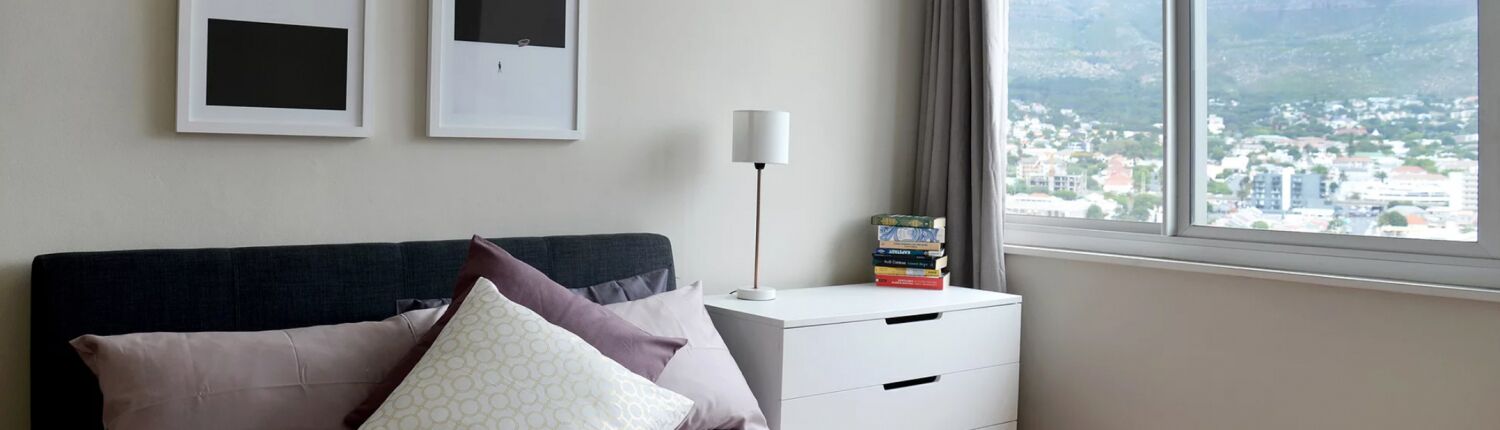 A bedroom with a bed, pillows, a bedside table with a lamp, and a window with a view of a cityscape.