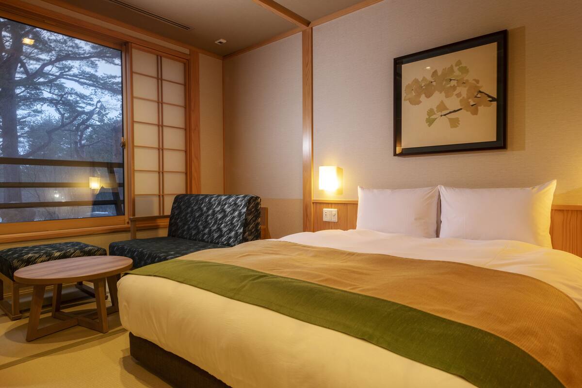A hotel room with a large bed, a small table, a chair, and a window with a view of trees.