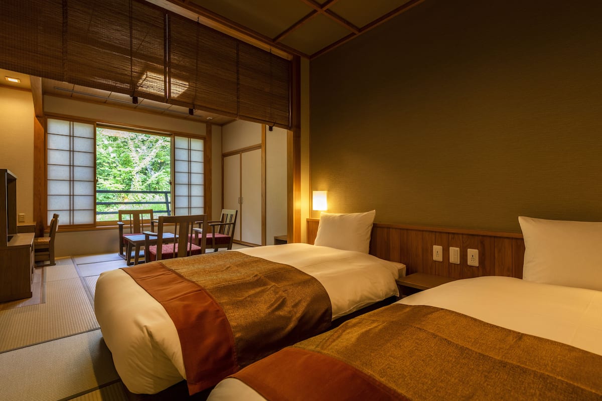 A traditional Japanese-style hotel room with tatami flooring, a low wooden bed, a desk, and a view of greenery through large windows.
