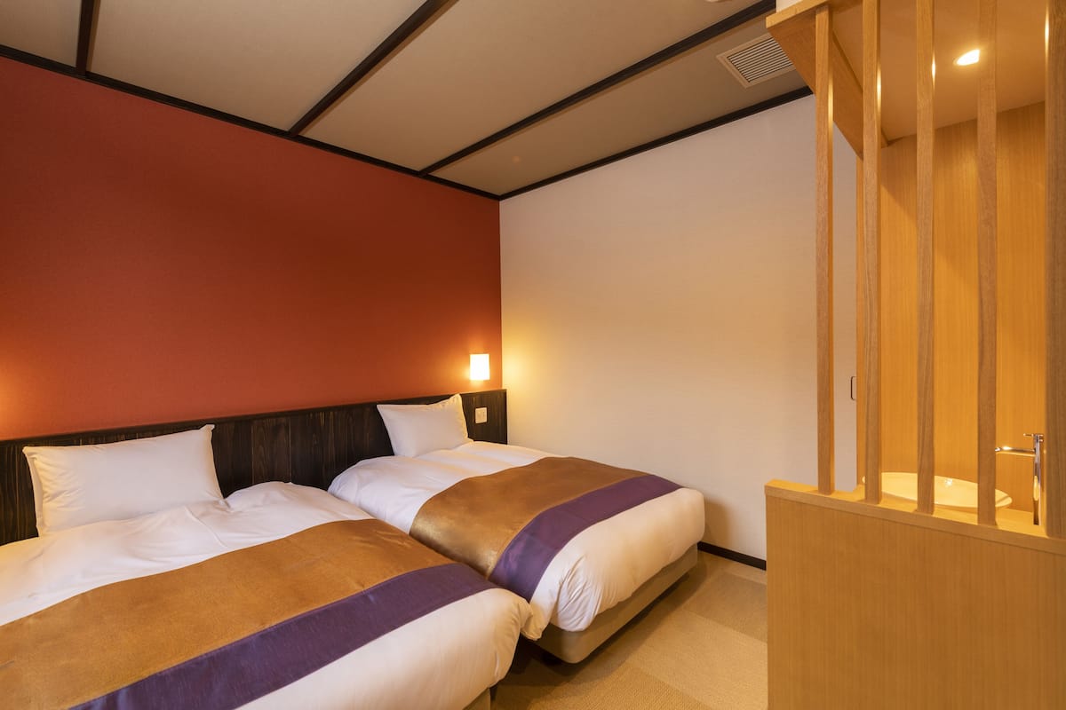 A hotel room with two beds, a wooden headboard, a nightstand, and a wooden partition with a sink.
