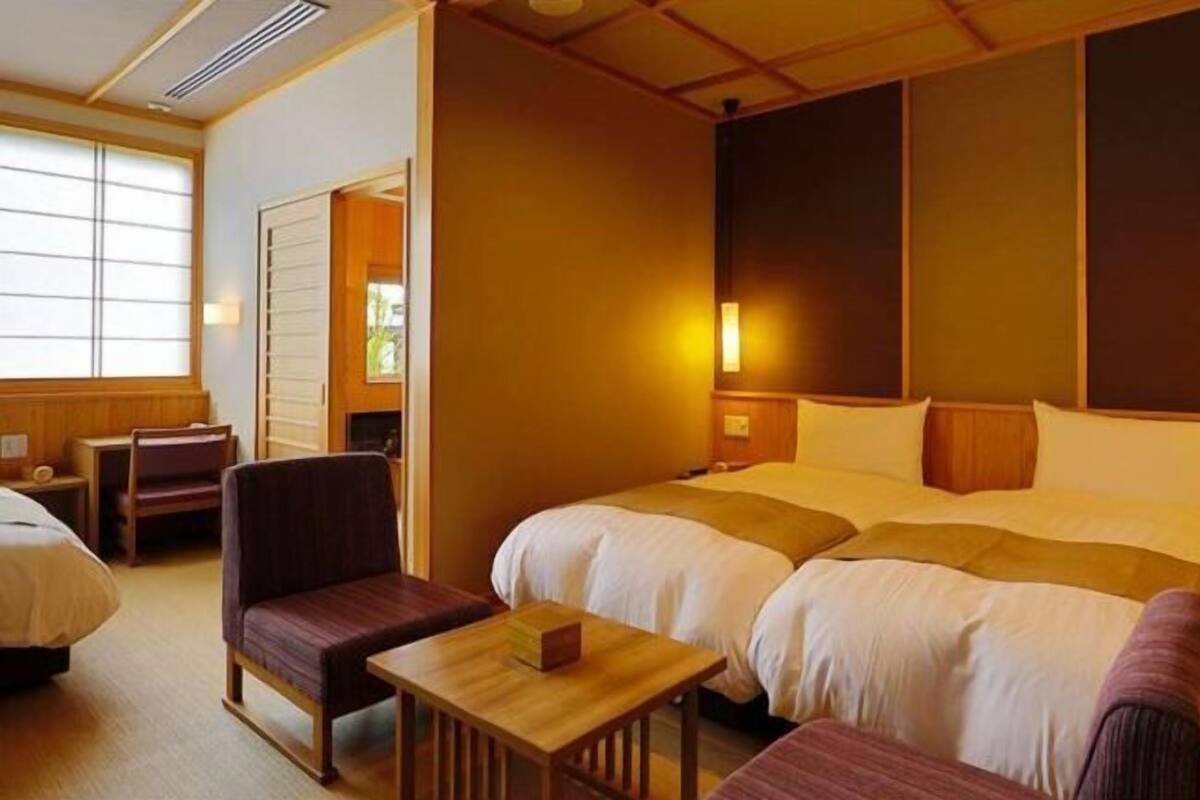 A hotel room with two beds, a chair, a small table, and a window.