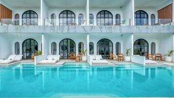A large, clear swimming pool with lounge chairs and potted plants, surrounded by a white building with arched windows and balconies.