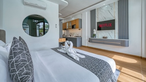 Ein modernes Hotelzimmer mit einem großen Bett, einem runden Spiegel, einer Küchenzeile und einem an der Wand befestigten Fernseher, der das Netflix-Logo zeigt.