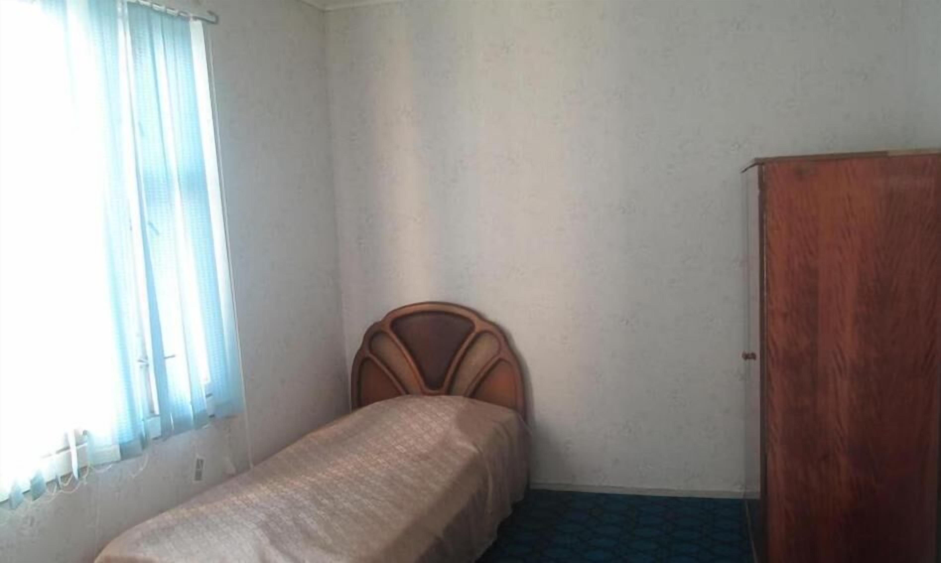 Ein kleines Zimmer mit einem Einzelbett, einem Holzkopfende, einem Kleiderschrank und einem Fenster mit Jalousien.