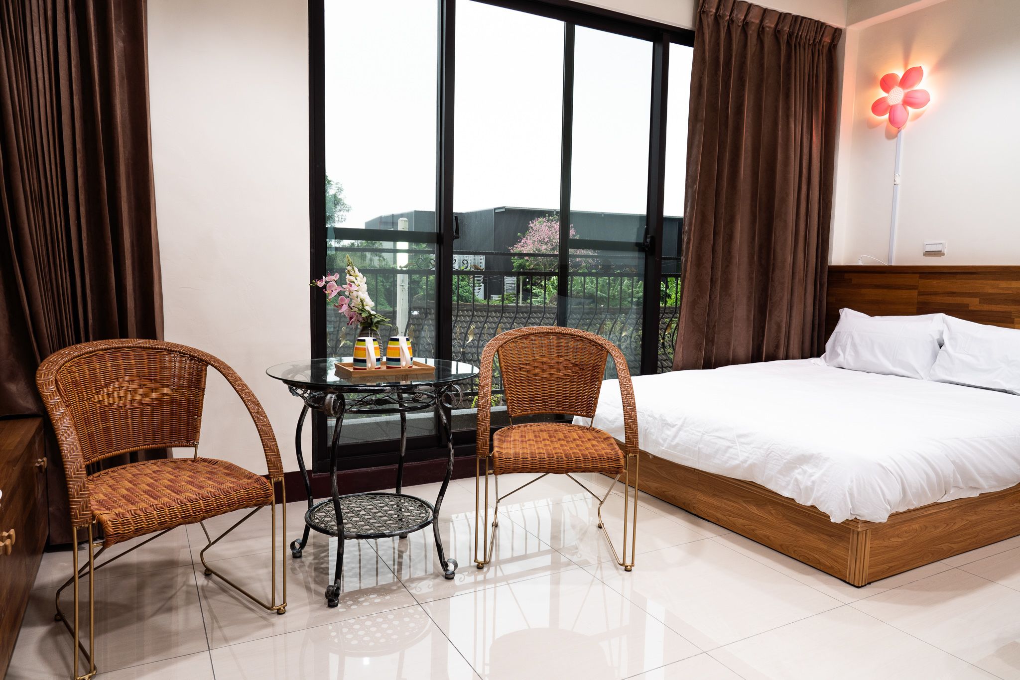 A hotel room with a large bed, two wicker chairs, a small table with a vase, and a view of greenery through large windows.