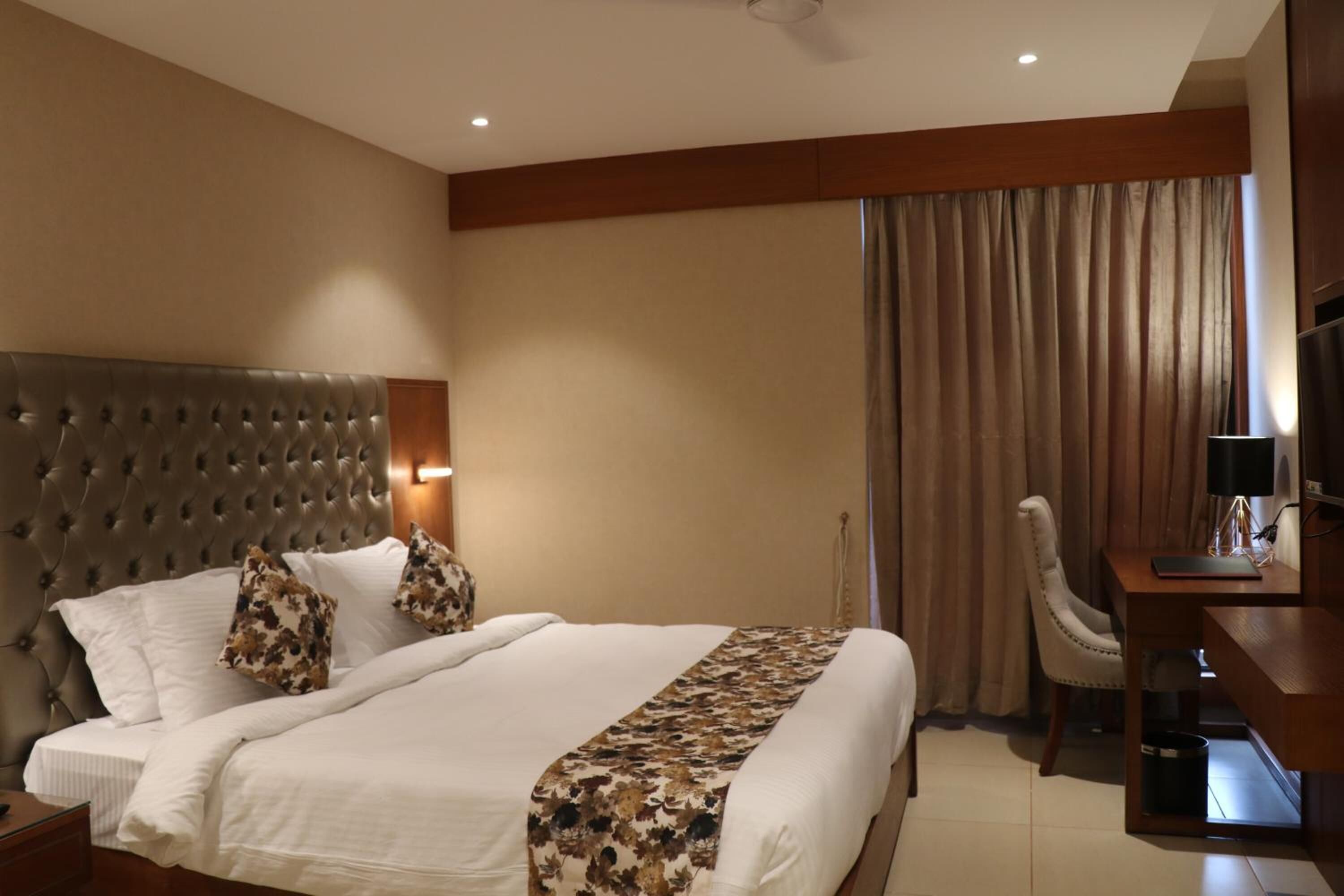 Una habitación de hotel con un cabecero capitonado, una cama grande con ropa blanca, un edredón estampado con motivos florales, un escritorio con lámpara y un televisor.