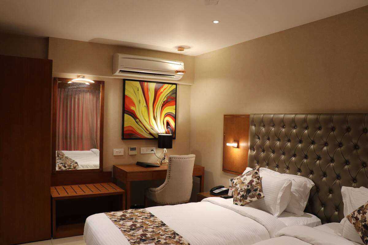 A hotel room with a large bed, a desk with a chair, a mirror, an air conditioner, and a colorful abstract painting on the wall.