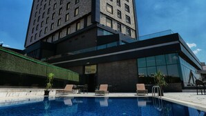 Ein modernes Hotel mit großzügiger Poolanlage, einer Glasfassade und einem hohen Gebäude im Hintergrund.