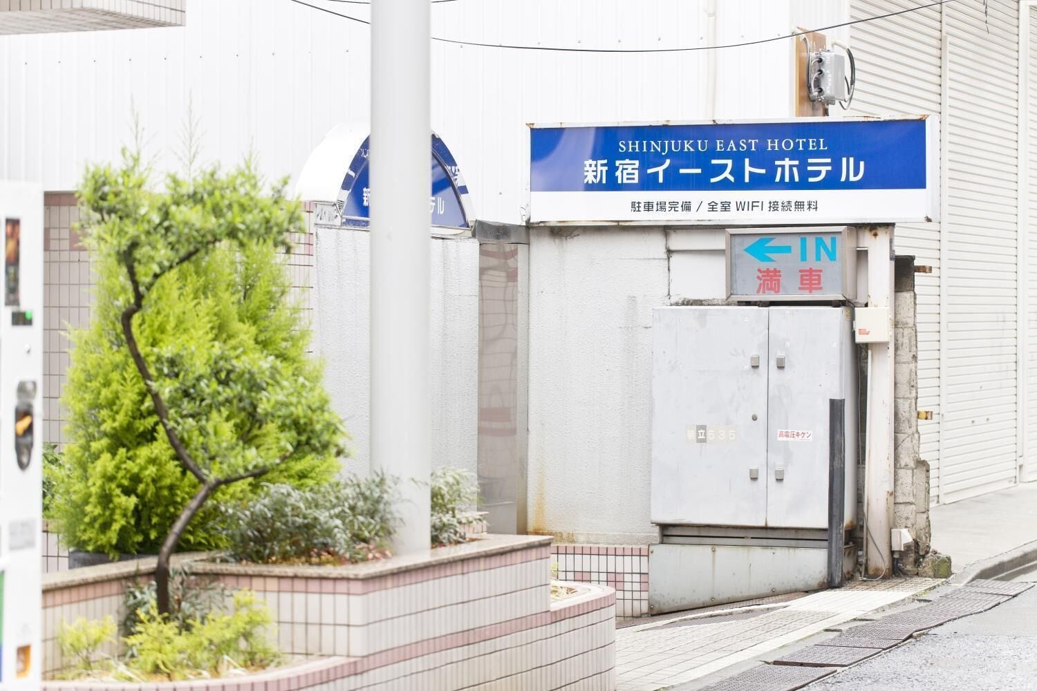 Entrada de un hotel con un letrero de Shinjuku East Hotel, una puerta con la inscripción "IN" y una caja de servicios.
