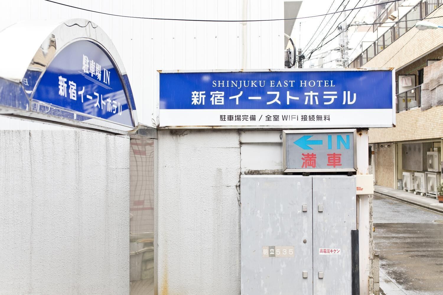 A hotel entrance with a sign for Shinjuku East Hotel, featuring a directional arrow and a contact number.