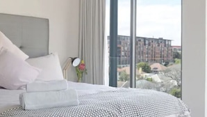 A modern hotel room with a large bed, a view of the city through floor-to-ceiling windows, and a cityscape visible outside. - Lily By Stay Amazing (Cape Town)