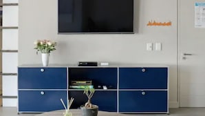 A room with a TV mounted on the wall, a blue cabinet with drawers, a vase with flowers, and a door with a sign. - Lily By Stay Amazing (Cape Town)