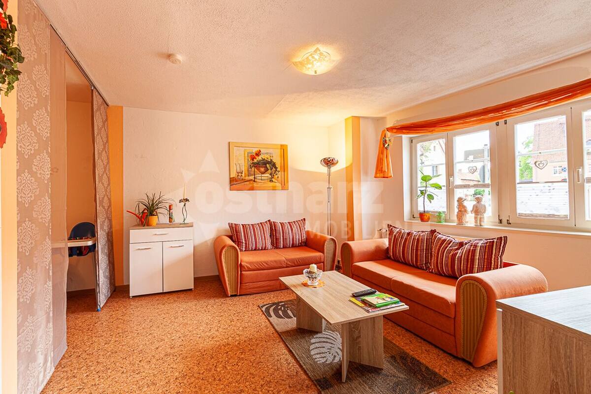 Ein gemütliches Wohnzimmer mit einer Sofagarnitur, einem Couchtisch und einem kleinen Schrank. Ein Fenster mit Vorhängen, eine Pflanze und ein Gemälde zieren die Wand.