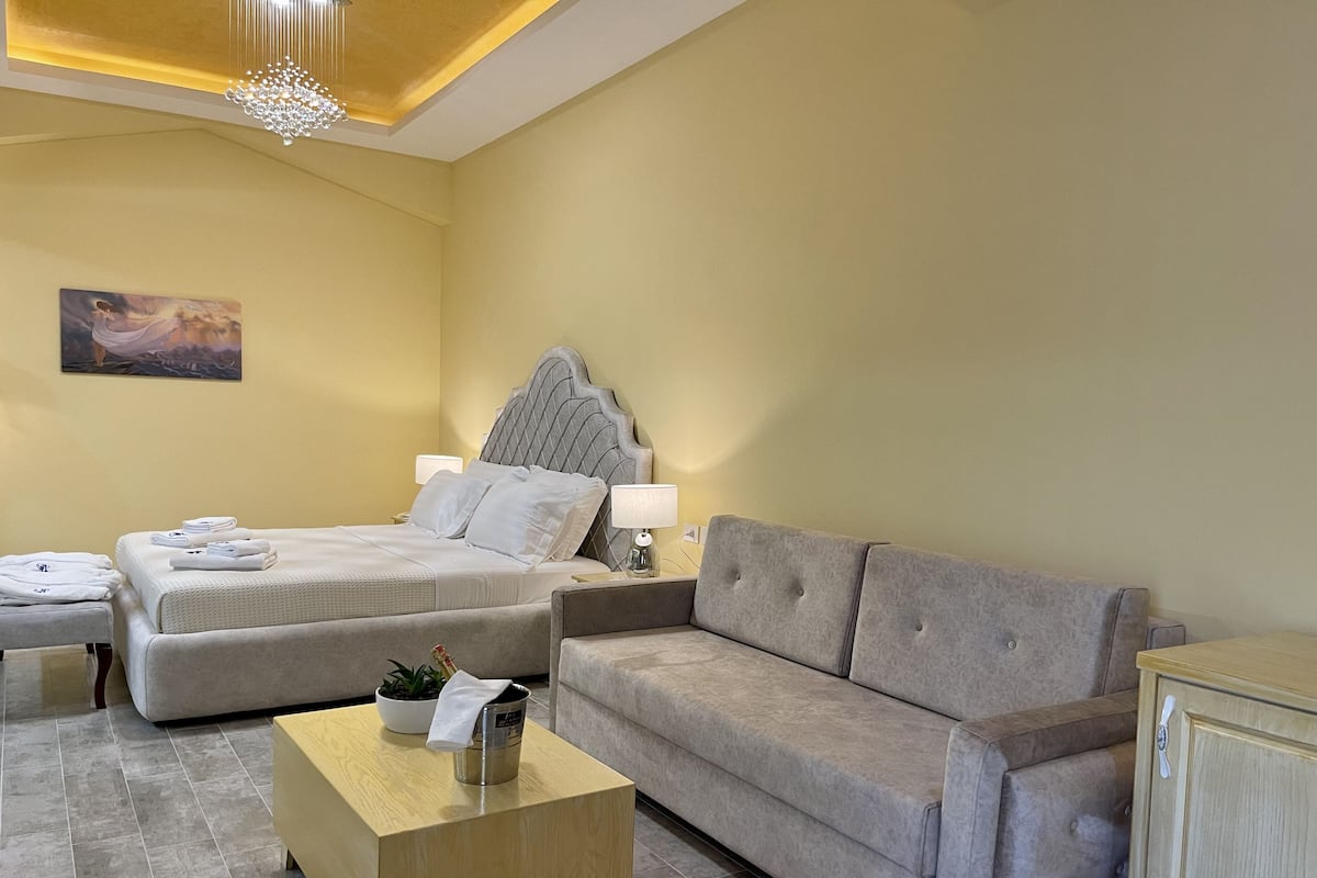 A hotel room with a bed, a sofa, a wooden side table, and a painting on the wall.