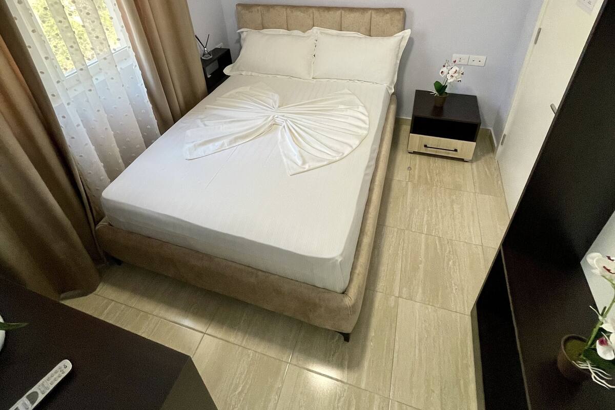 A neatly made bed with a decorative bow on the bedspread, a bedside table with a plant, and a television on a stand.