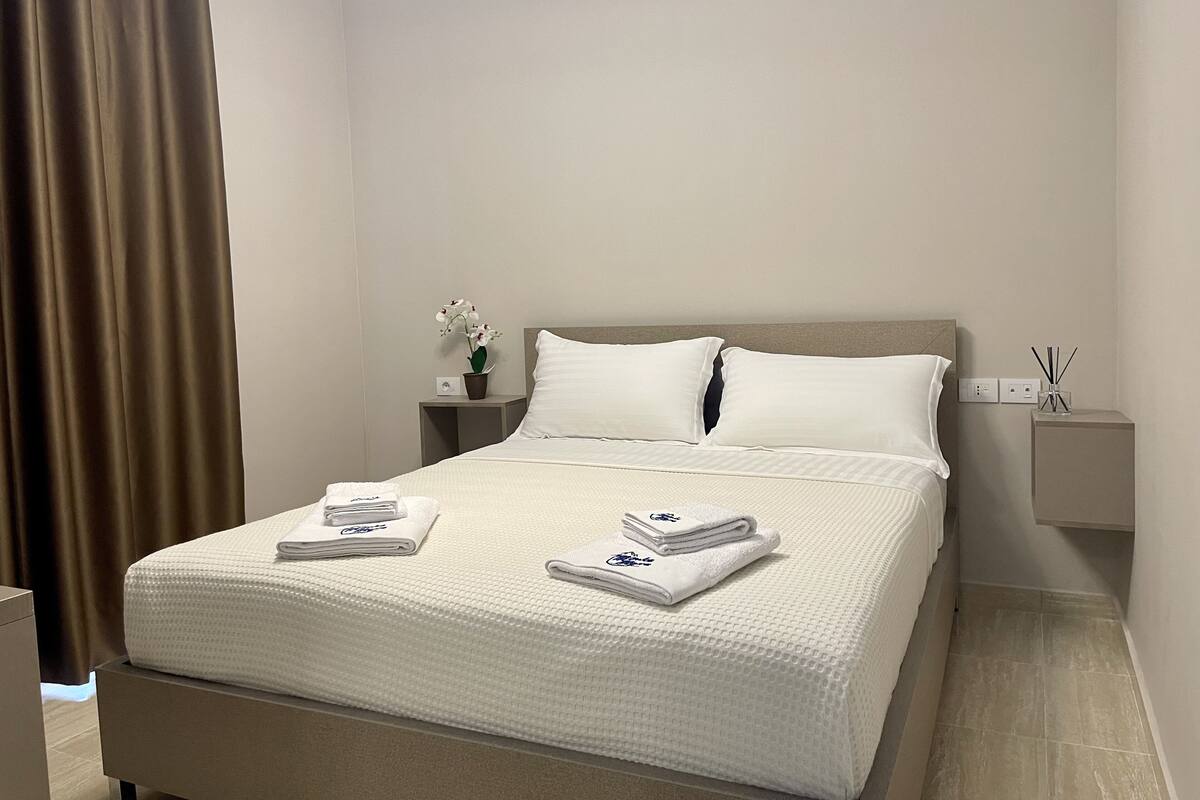 A neatly made bed with white bedding and towels, a bedside table with a small plant, and a nightstand with a lamp.