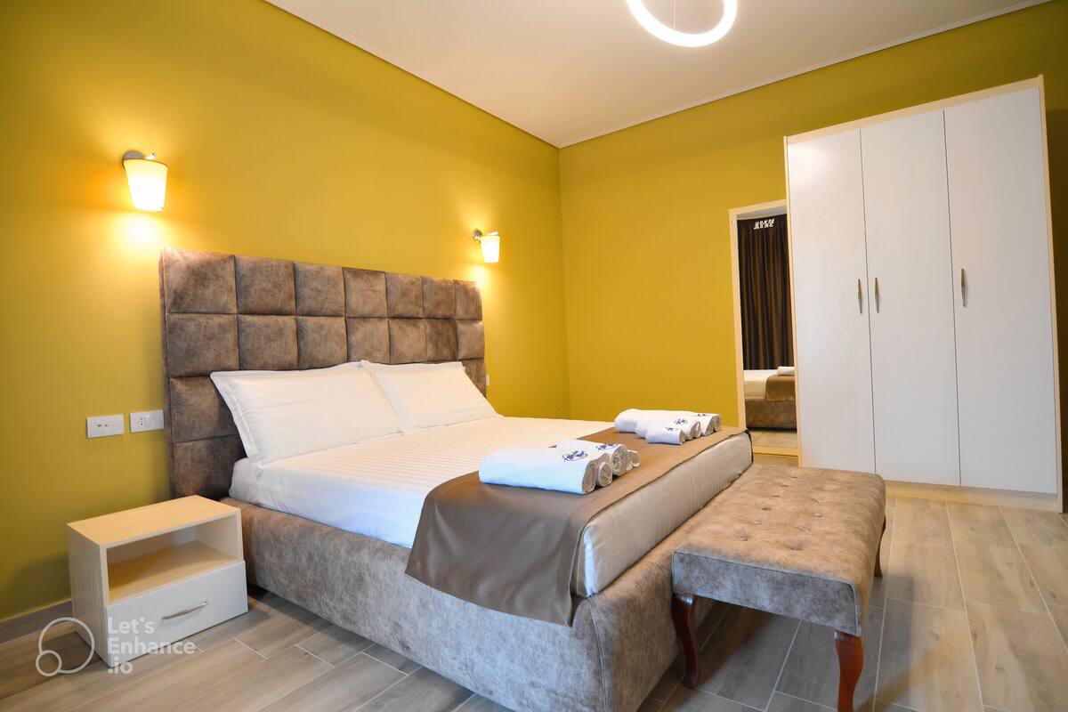 A bedroom with a bed, bedside table, bench, wardrobe, and wall-mounted lights.