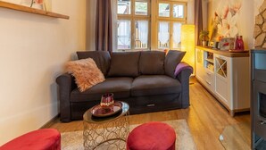 A cozy living room with a dark sofa, a small round table with a drink, and a red ottoman. There are large windows with curtains, a lamp, and a painting on the wall. - Apartment unter dem Schloss (Quedlinburg)