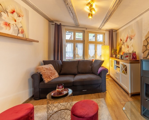 A cozy living room with a dark sofa, a small round table with a drink, and a red ottoman. There are large windows with curtains, a lamp, and a painting on the wall.
