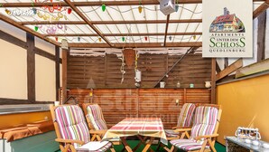 An outdoor patio with a wooden table and striped chairs, a hanging light fixture, and a wooden wall. - Apartment unter dem Schloss (Quedlinburg)