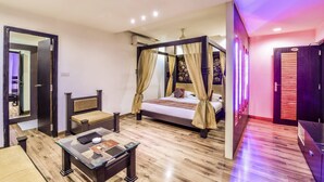 A bedroom with a four-poster bed, a sitting area with a sofa and armchair, a small table with a book and a remote control, and a wardrobe with a mirror. - Raghu Mahal Hotel (Udaipur)